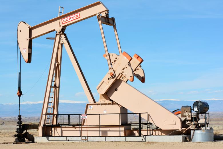 A large, tan-colored pumpjack stands in a flat, arid landscape under a clear blue sky. On the right, large counterweights with orange accents are attached to the crank arms. In the background, low-lying desert terrain transitions into a distant range of snow-capped mountains beneath a bright horizon.