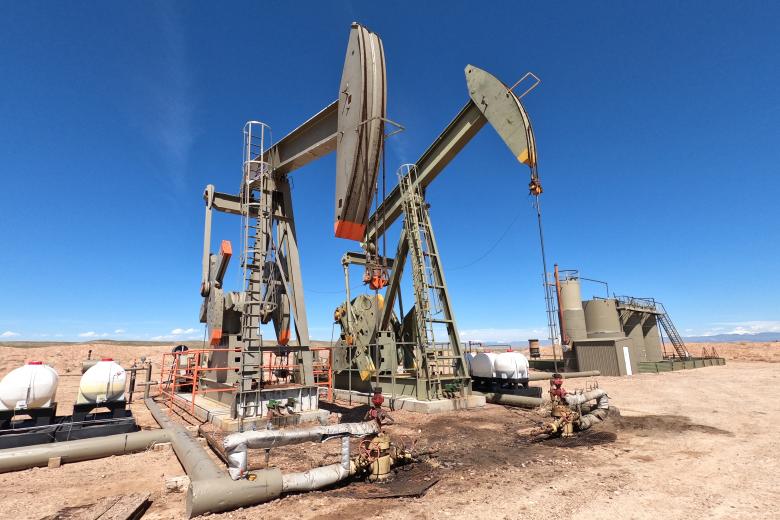 Two large, olive-green industrial pumpjacks stand in a flat, arid desert landscape under a clear, bright blue sky. In the foreground and mid-ground, various storage tanks, white pressurized vessels, and insulated piping systems are visible on the sandy ground.