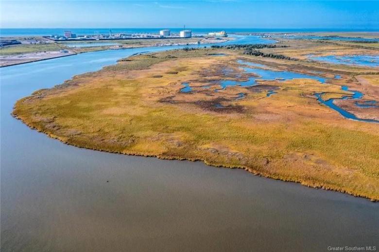 Aerial view of grassy marshland bordered by calm water, with industrial tanks and buildings visible in the distance under a clear blue sky.