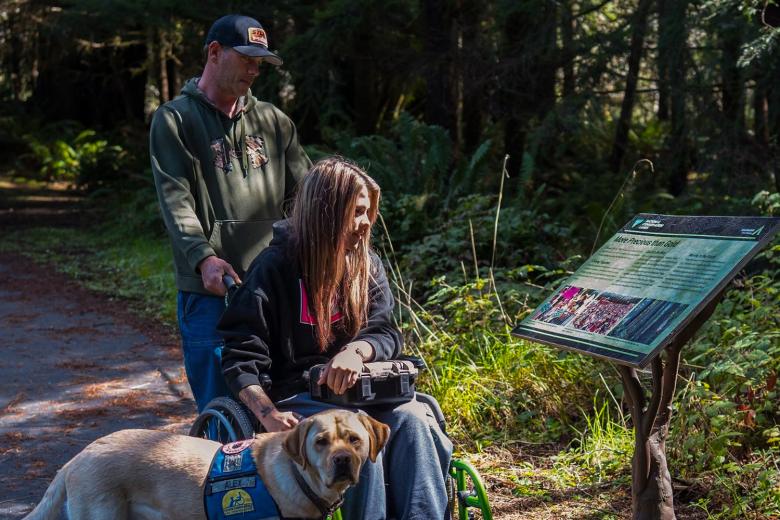 A person in a wheelchair on a forest trail, accompanied by another person and a service dog pausing to look at a BLM interpretive sign. 