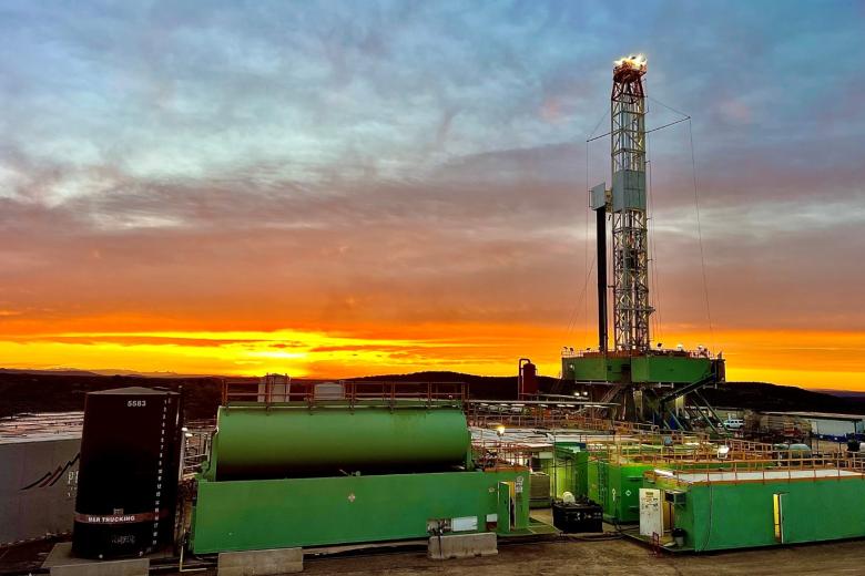 An onshore oil and gas drilling rig stands against a vibrant orange and blue sunrise sky. In the foreground, large green industrial mud tanks and a vertical black storage tank sit on a dirt lot. The tall metal derrick tower rises on the right side of the frame with its working lights illuminated.