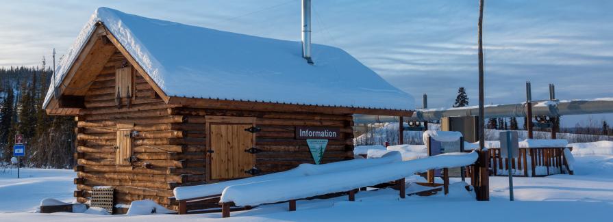 Snow blankets a log cabin with an "Information" sign. Pipeline in background