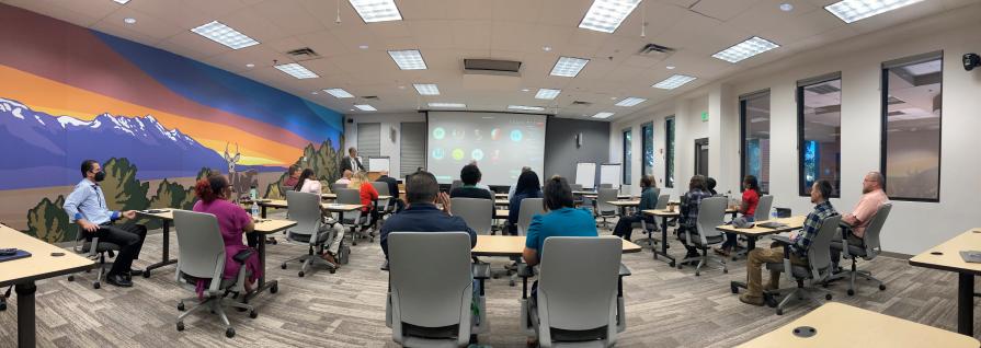 Group of people sitting in chairs behind tables listening to presentation in a National Training Center classroom with a large mural on the wall of a southwest landscape featuring a deer.