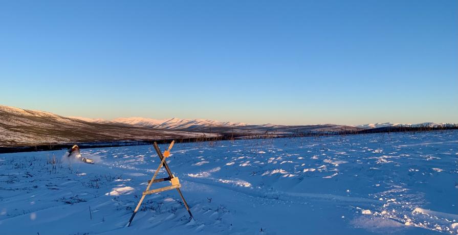 A snomobile rider kicks up snow as he rides past a tripod trail marker used in open areas. The mountains are lit by the setting sun.