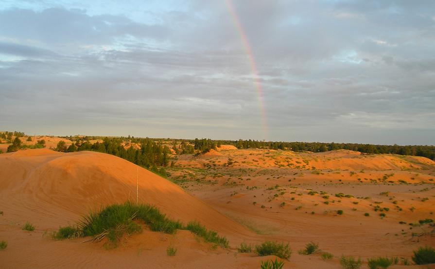 Sand dunes are lit up by post-rain shower sun. A rainbow arches overhead.