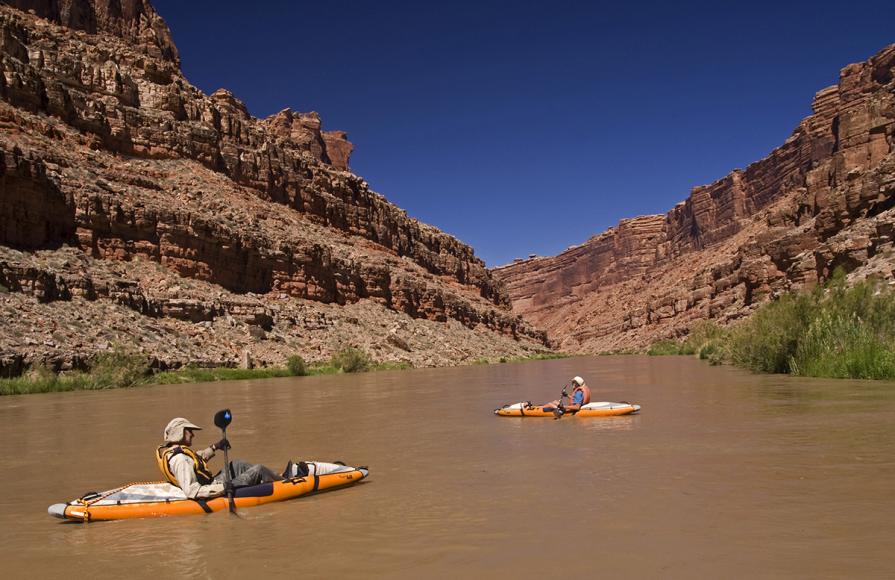 Two kayakers paddle over flat, brown water surrounded by sheer red cliffs. Photo by Sue Cullumber and the Share the Experience Photo Contest.