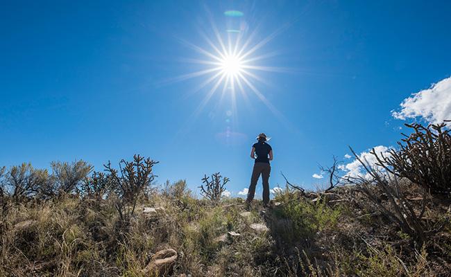 A woman stands in a high desert landscape with a sunburst overhead.