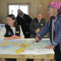 Native elder woman standing and pointing to map of the Bering Sea-Western Interior Planning are map with BLM Planning staff nearby during a public meeting
