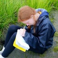 Student writing in journal while sitting on the ground near tall grass.