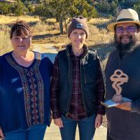 Left to right: Chief of Interpretation Lisa Dittman, El Malpais National Park; Kymm Gresset, Rio Puerco Field Office Manager; and Sculptor Walter Torres, stand smiling. Walter is holding a scaled down version of the sculpture. There are trees in the background. 