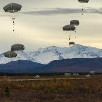 Paratroopers with open parachutes float through the sky and land on the grassy plain with snow-capped mountains in the background.