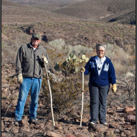 Two hikers with stand in a rocky desert landscape with cacti and mountains in the background