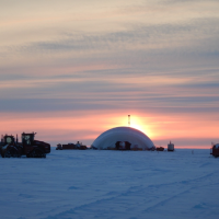 Snow-covered ground, with an orange sunset behind a large inflatable dome. Heavy equipment nearby.
