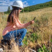 Woman squatting in tall grass with a hard helmet on. 