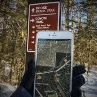 person holding smart phone with georeferenced map opened up in mobile app with trail and trail signage in the background during winter at Campbell Tract