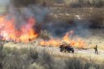 a fire burns through vegetation as firefighters look on