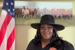 Woman with hat in front of horse picture and near an American flag. 