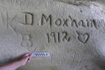 A person holds a measuring stick up against a historic inscription that is on a rock face. 