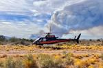 Helicopter sits stationary in a desert lands scape with short vegetation in the area (helispot). A large plume of smoke is visible behind the helicopter in the distance. 