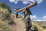 MCC crew members carrying timber over their shoulders down the trail on the side of a grassy hill. 