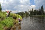 A person stands on a green vegetative river bank while another wades in the water to measure the depth of the water.