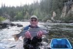 A man is standing in a river holding a large fish and smiling. Next to him is a blue bucket. 