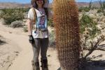 A woman, smiling, in a sun hat and snake gaiters, stands next to a large cactus. She is surrounded by green shrubbery and gray mountains in the background. 