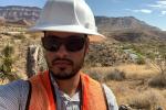 Carlos Joya, BLM Arizona's state engineer, takes a selfie. He is wearing a white hardhat, black glasses, and an orange high-visibility vest. The Virgin River Campground area is behind him.