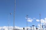 A person stands looking up at 30-foot metal poles with weather vanes on the top supported by tripod legs. 