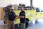 two people shake hands in front of a yellow fire engine. One person hands keys to the other.