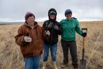 Three people wearing cold weather gear smile at the camera. One of them holds a shovel and the other two hold seedlings.