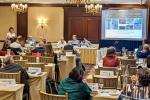 People sitting a various tables looking at a large screen while listening to a presenter