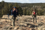 Two caucasian men pose outdoors in hiking clothes carrying metal stakes and wheels of wire on their backs.