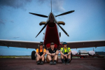 Three Caucasian women sit on the ground in front of a small airplane
