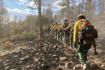 A line of people in green pants, yellow shirts, black backpacks, and hard hats walks through a burned forest.