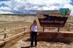 A woman in sunglasses stands facing the camera and smiling next to a sign with the BLM logo. The sign reads “Gunnison Gorge National Conservation Area.” Behind her, beyond an area of shrubland, are light brown and green mountains beneath a sky with large white clouds and patches of blue. 