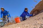 BLM employee Ralph Burrillo applies a fresh coat of mud to the adobe wall while BLM employee Theresa Condo hauls more mud. They are both wearing hats and blue long sleeve shirts.