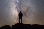 A photo showing the silhouette of a man standing on a high point of ground looking up at the night sky, which is a blend of gray and beige, dotted heavily with stars. The Milky Way is positioned vertically in the center of the photo.  