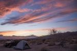 A jeep is parked next to a tent on a desert landscape, with a mountain in the background and the setting sun turning the sky and clouds pink and blue.