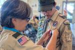 A Boy Scout in uniform looks down as an adult leader pins the Eagle Scout medal to his chest. The scout wears a sash covered in merit badges, and other scouts are visible seated in the background.