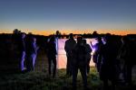 A group of people gathers around a brightly lit screen in a field at dusk, with a colorful sunset in the background.