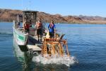 Workers in life jackets unload an orange metal frame holding a bundle of brush into a lake, with rocky shoreline terrain visible in the background.