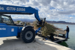 A blue telehandler forklift loads pine trees onto a barge at a boatlanding on a lake.