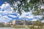 White limestone cliffs rise above a calm river under a bright blue sky with scattered clouds, framed by green leafy branches.