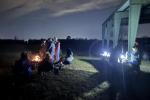 A group of people gathered around a campfire at night, with some sitting in chairs and others preparing food under a shelter.