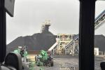 View from a vehicle showing heavy machinery and large coal piles at an industrial site, with conveyor belts and cloudy sky.