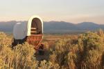 A covered wagon surrounded by sage brush with distant mountains at sunset