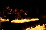 Musicians playing string instruments on a dark stage surrounded by numerous glowing candles, creating a warm, intimate atmosphere.