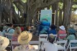 Artist painting a marine scene on a large canvas outdoors, with an audience seated on white chairs under large tree branches.