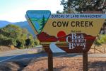 A triangular sign reading “Bureau of Land Management Cow Creek National Back Country Byway” sits in a grassy area near a two-lane road bordered by green trees, with tree-covered hills in the distance. The marker includes both the BLM and National Back Country Byway logos. 